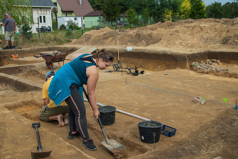 osoba używająca łopaty na tle stanowiska archeologicznego